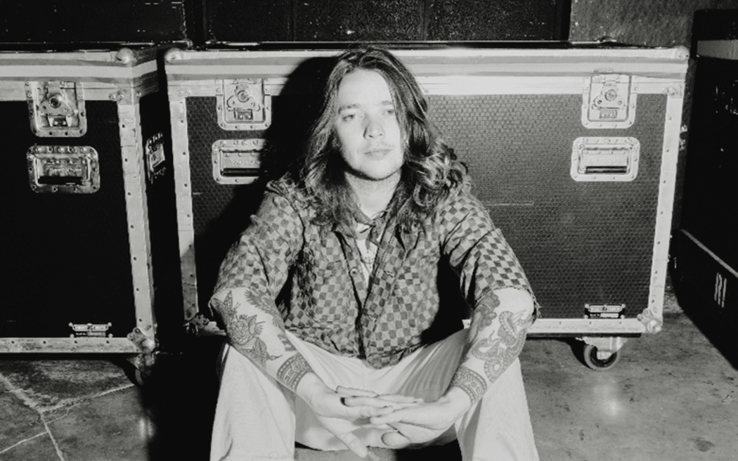 This is a black and white photo of Billy Strings. He is sat in front of a large flight case. It appears to have been taken backstage at a gig, but could also have been staged to give that impression. He has long hair flowing over his shoulders, and you can see his socks peaking out over the top of a pair of hith top Chucks.