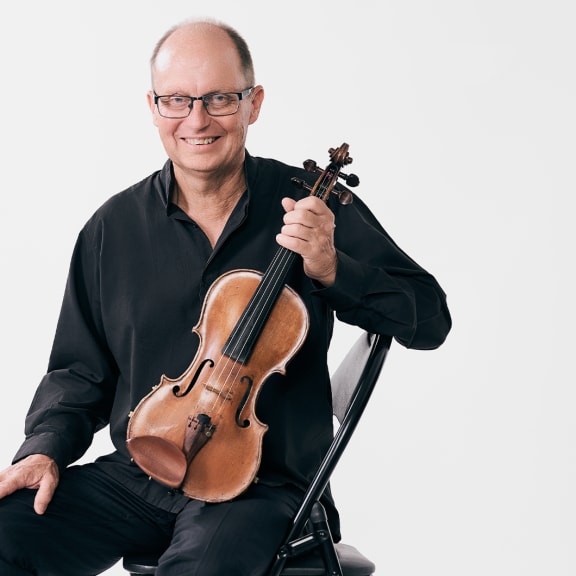 NZSO Associate Concertmaster Donald Armstrong poses on a black chair set against a white background. Donald is dressed in black shirt and trousers, wears glasses and is holding a violin.
