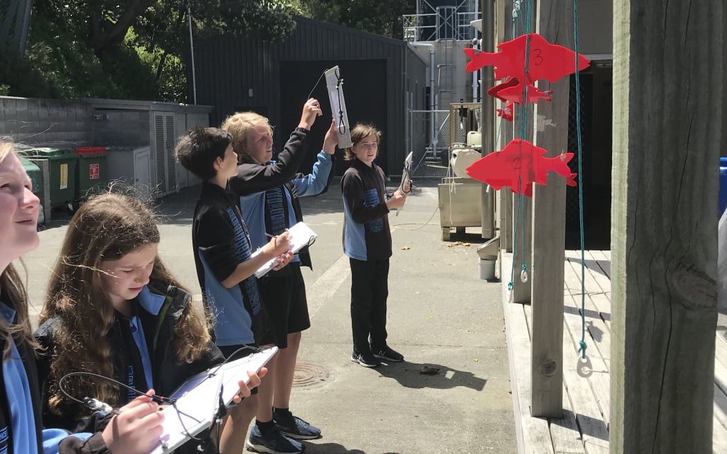 Students stand outside in front of a washing line with red fish outlines hanging from them. They have white slates on to which they are trying to estimate fish sizes.