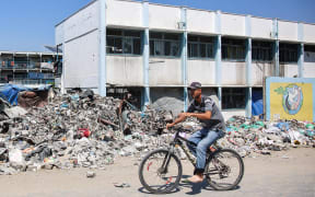 A man cycles past trash and rubble outside a school run by the UN Relief and Works Agency for Palestine Refugees (UNRWA) being used as a makeshift shelter for Palestinians displaced by conflict in the Jabalia camp for Palestinian refugees in the northern Gaza Strip on August 14, 2024 amid the ongoing conflict in the Palestinian territory between Israel and Hamas. (Photo by Omar AL-QATTAA / AFP)