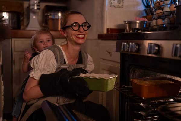 Simone Collins, with a toddler strapped to her back, holds a cake she's taken out of the oven whilst wearing oven mitts.