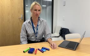 A woman wearing a striped shirt sits at a desk with six different colourful vapes laid out in front of her. She has a serious look and also has a laptop.