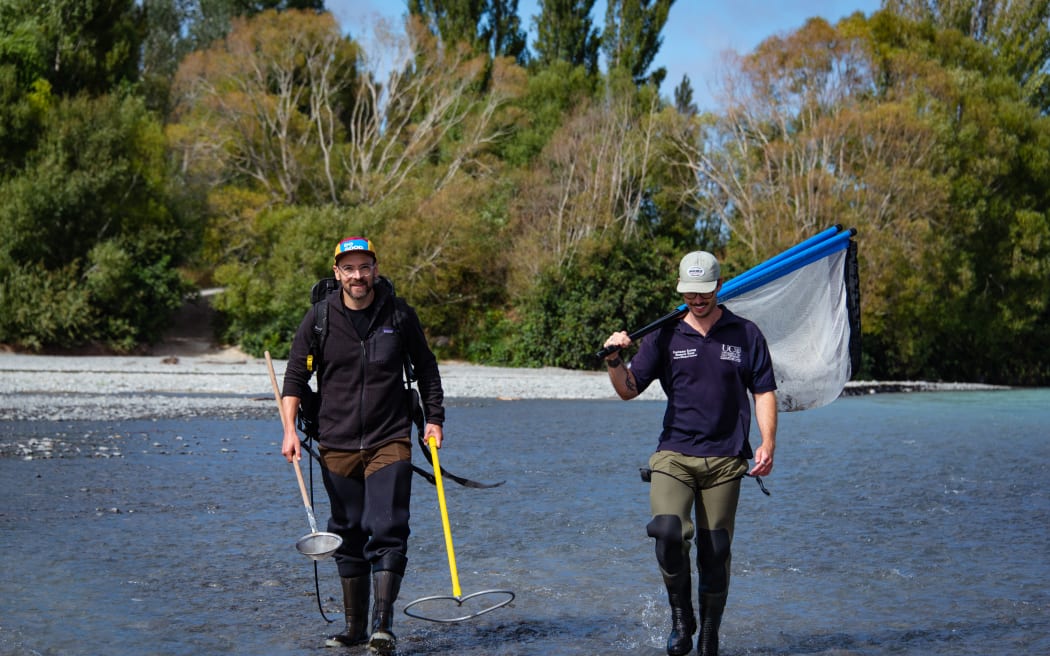 Two men walk across a shallow braided river. One if holding a net over his shoulder, the other is holding two scoop-like contraptions.