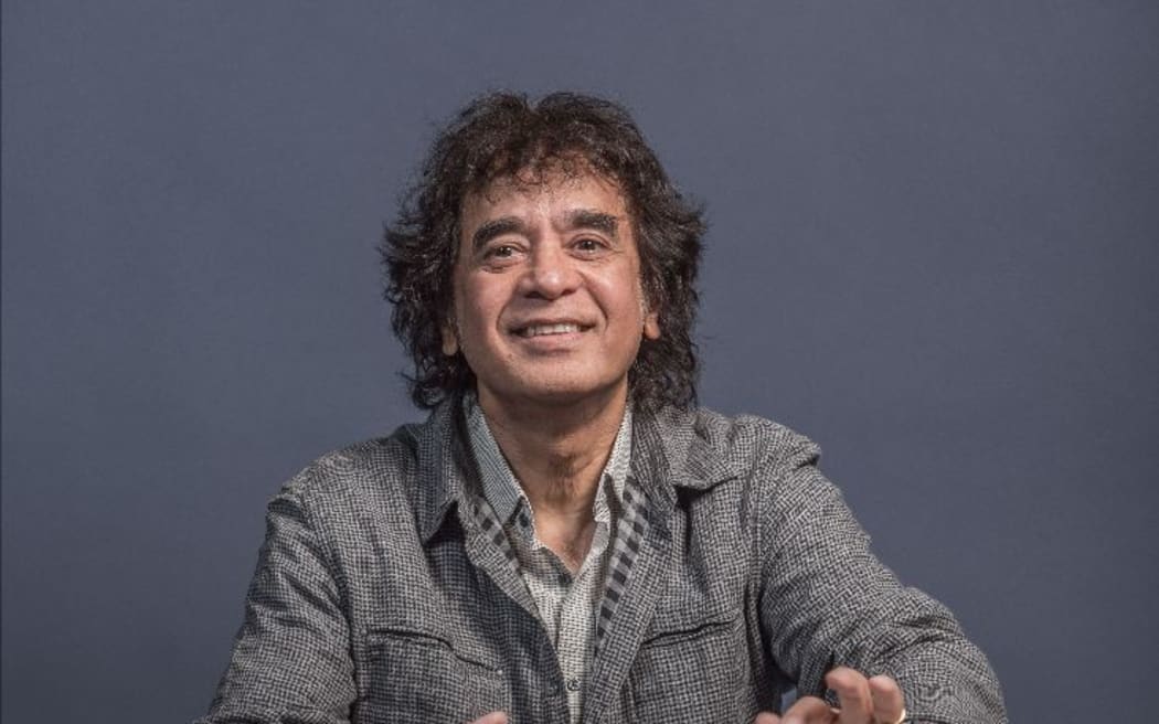 Zakir Hussain sitting cross-legged playing the tabla in grey clothing.
