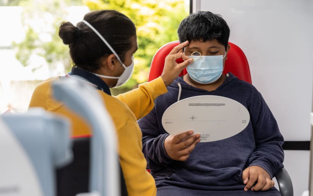 A boy sits in a patient's chair receiving an eye exam. The optometrist holds a lens to his eye while he reads from a chart.