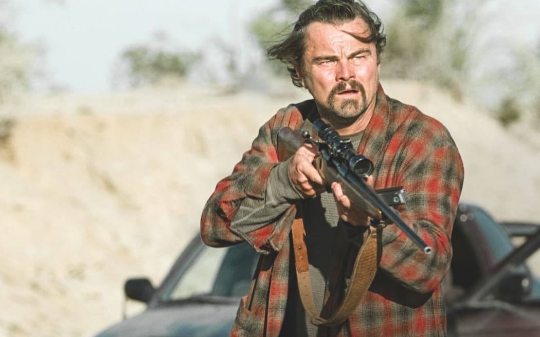 Still from the 2025 film One Battle After Another showing star Leonardo Di Caprio wielding a rifle while wearing a faded housecoat.