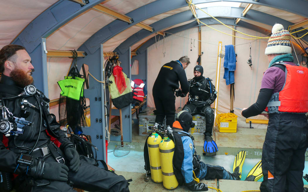 Inside a tent with metal beams, a person in full scuba gear and drysuit with head cover sits on the edge of a hole in the ice. A second person in scuba gear sits behind the hole, with a man attending to them. A third person in scuba gear sits on the left, he has a beard and is not wearing a mask or head cover. A person wearing a red lifejacket and thick beanie watches with a clipboard from the right.