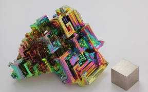 The chemical element bismuth as a synthetic crystal. The iridescent surface is a very thin layer of oxidation. Beside it is a high purity (99.99 %) 1 cm3 cube of bismuth for comparison.