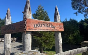 The entryway to Re-Engineered Ironbark, showing four large upright wooden poles capped with steel with a large wooden sign. In the foreground, a gateway made of riverstones