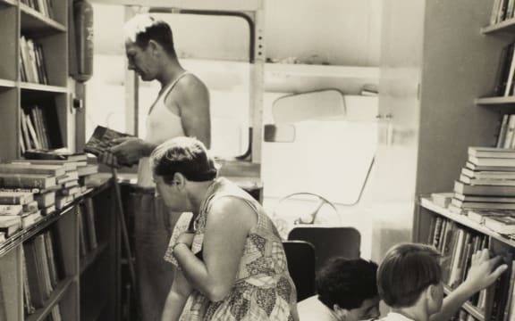 View of three women and a man as seen inside the van that houses the portable library service, shelves of books and a pile of books on a table are visible at left and at right.
