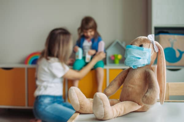 A woman plays with a child in a play room and in the foreground is a rabbit soft toy wearing a medical mask.