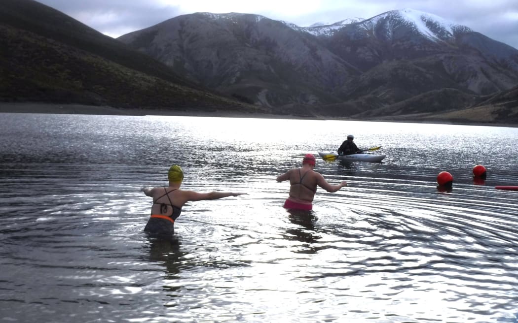Two swimmers dive into cold lake water, with a kayaker keeping waych from a distance. Mountains at the back of the lake are capped with snow.
