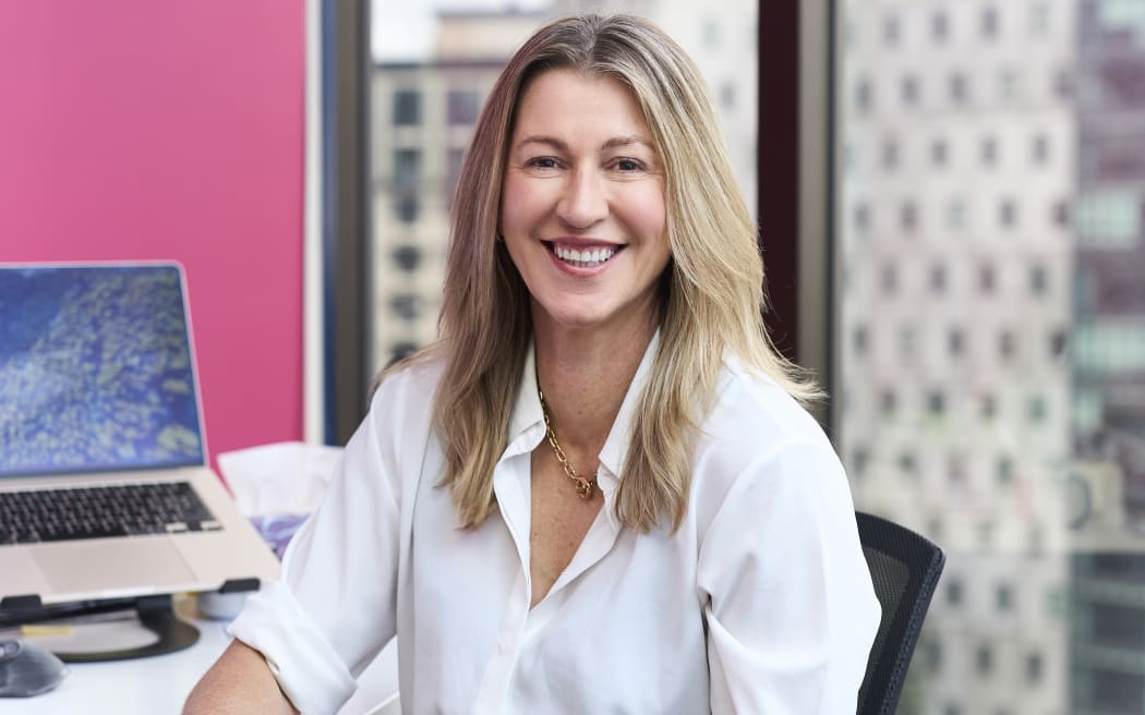 Woman with long blond hair and white shirt sits at desk smiling