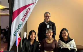 A group of people sit at an information kiosk, next to a banner that says BANYUHAY AOTEAROA. They all smile.