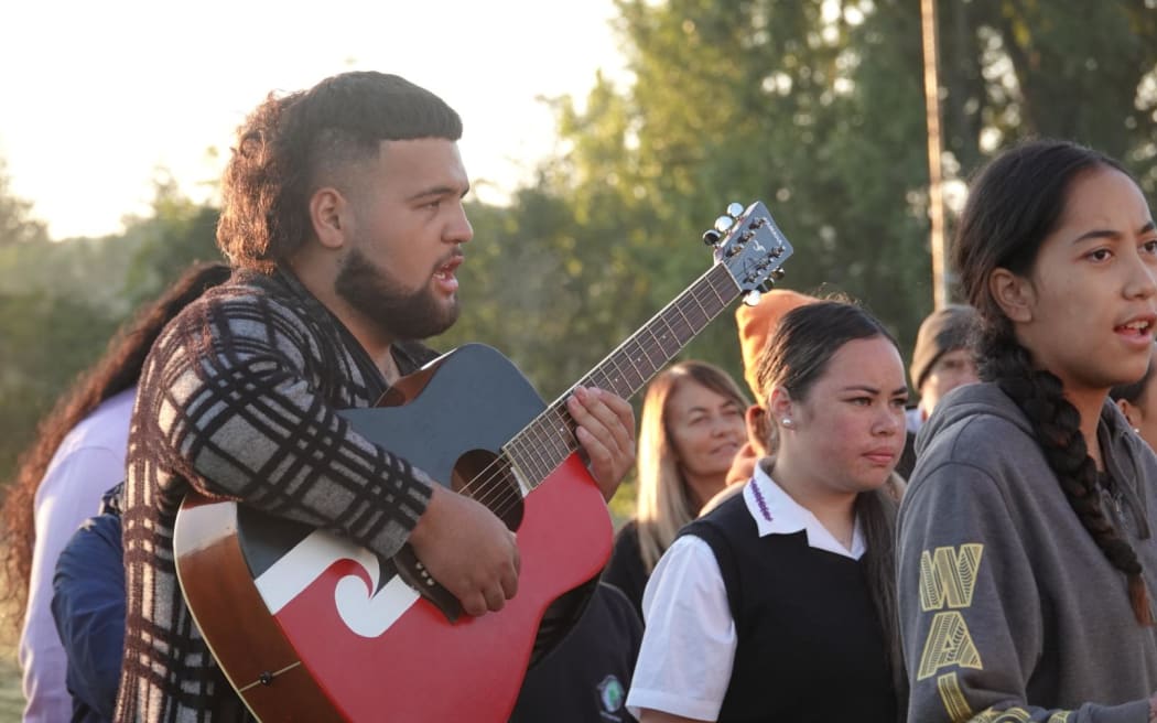 A person holds a guitar with a tino rangatiratanga flag painted on it while people next to them sing.
