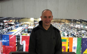 Ben Kepes wears a black hoodie and stands on an elevated platform. Behind him is the factory floor of Albion Clothing, with rows of workbenches and tools. There is a string of flags hanging over the factory floor.