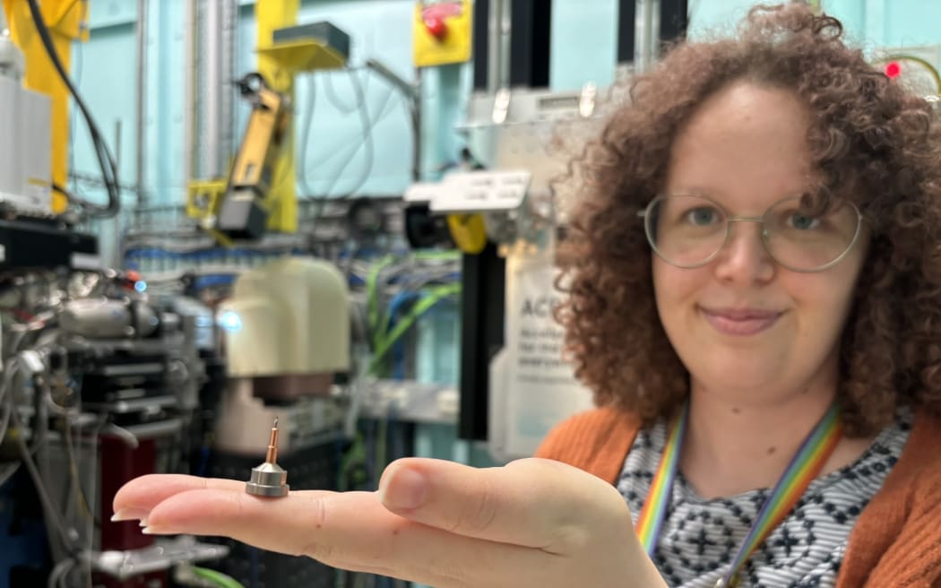 A woman with curly hair and glasses holds up a thimble-sized metal pin on her finger. She is standing in front of complicated looking machinery.