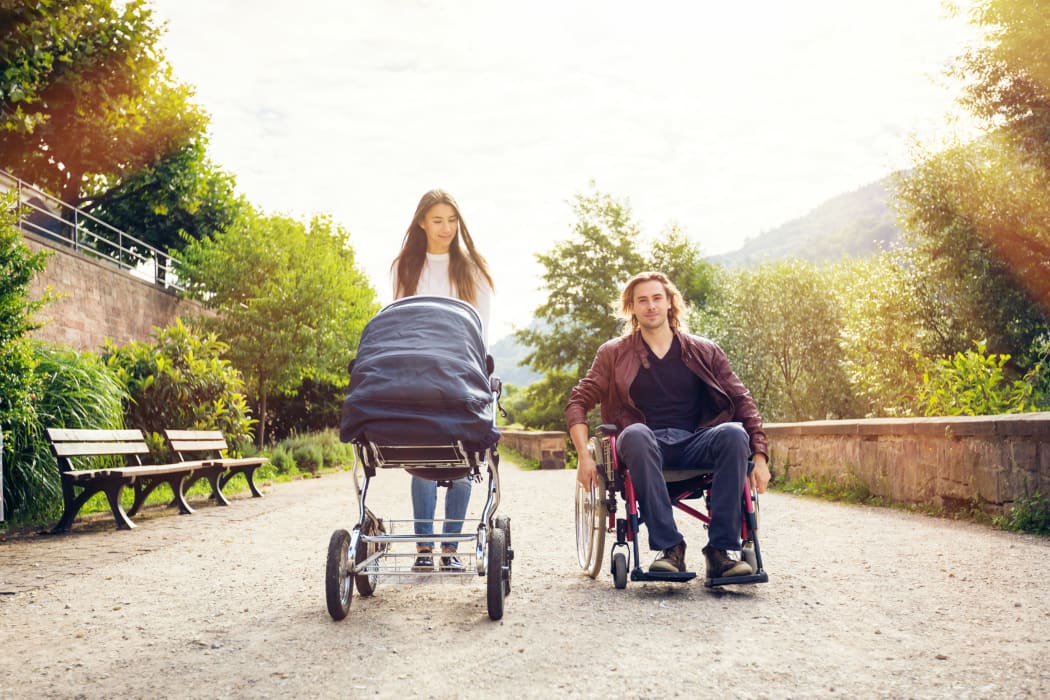 A photo of young parents enjoying time outdoors, taking their baby to a park. The father is sitting in a wheelchair.