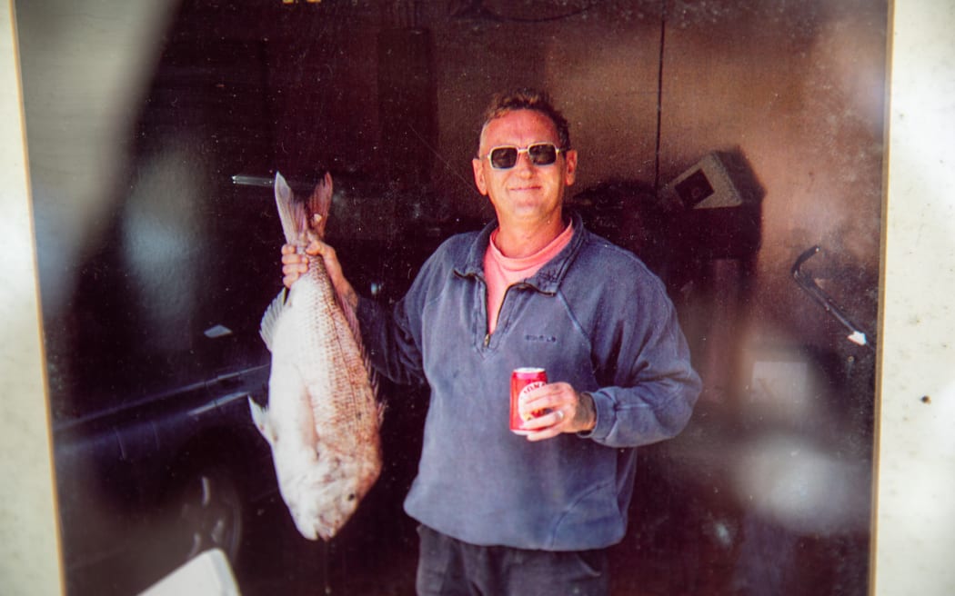 Archival photograph of a man (Ross Appelgren) holding a fresh caught fish in one hand and a can of beer in the other. He wears sunglasses and a blue sweatshirt and black shorts and smiles softly while looking at the camera.
