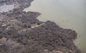 A P-3K2 Orion aircraft flies over an area of Tonga that shows Ash on homes and surrounding vegetation.