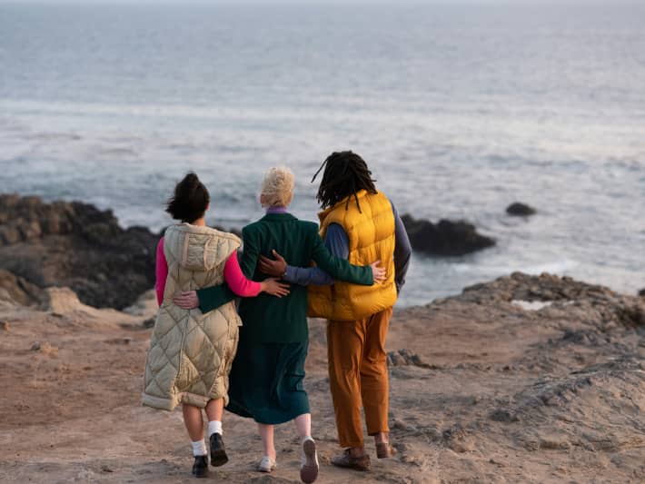 Three people embrace on a rocky coastline