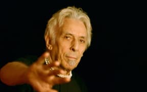John Cale wearing a necklace and raising his hand towards the camera