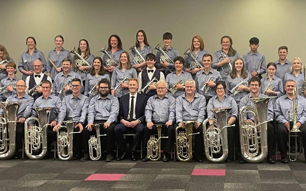 The Rolleston Brass pose for a band photo, seated in rows inside a large room. They all wear blue shirts and hold their instruments, smiling.