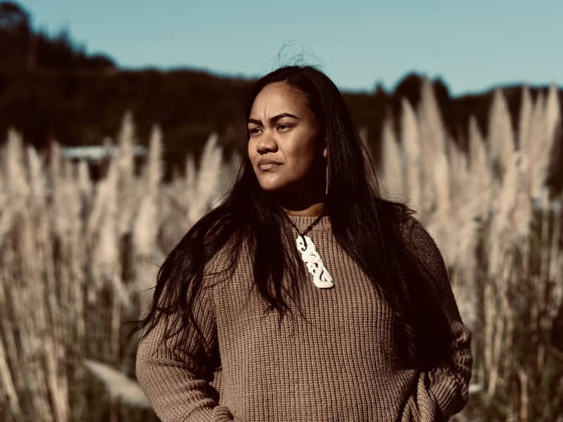 A Māori woman with long hair wears a carving around her neck and stands in a field.