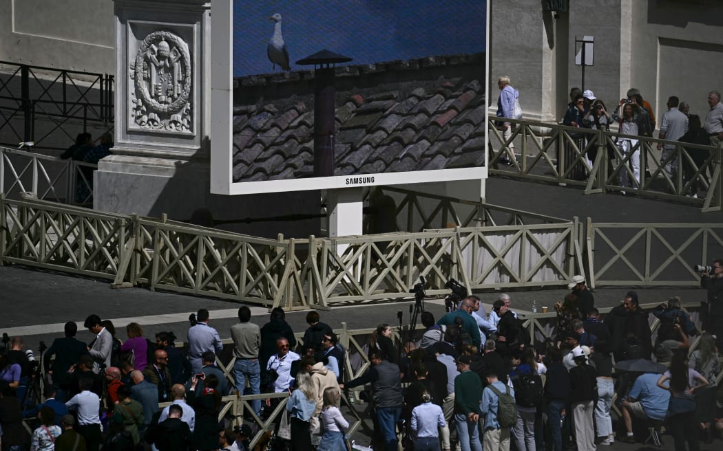 People watch a screen displaying a view of the chimney of the Sistine Chapel with a seagull at St Peter's Square on the second day of the conclave, in the Vatican on 8 May, 2025.