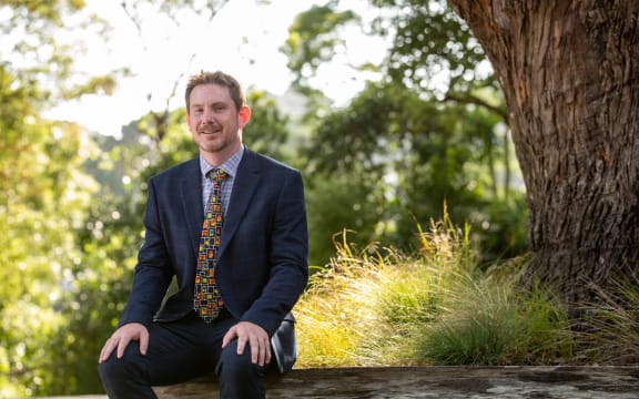 A man sitting in front of tussocks and trees. He is wearing a suit and tie and smiling at the camera.