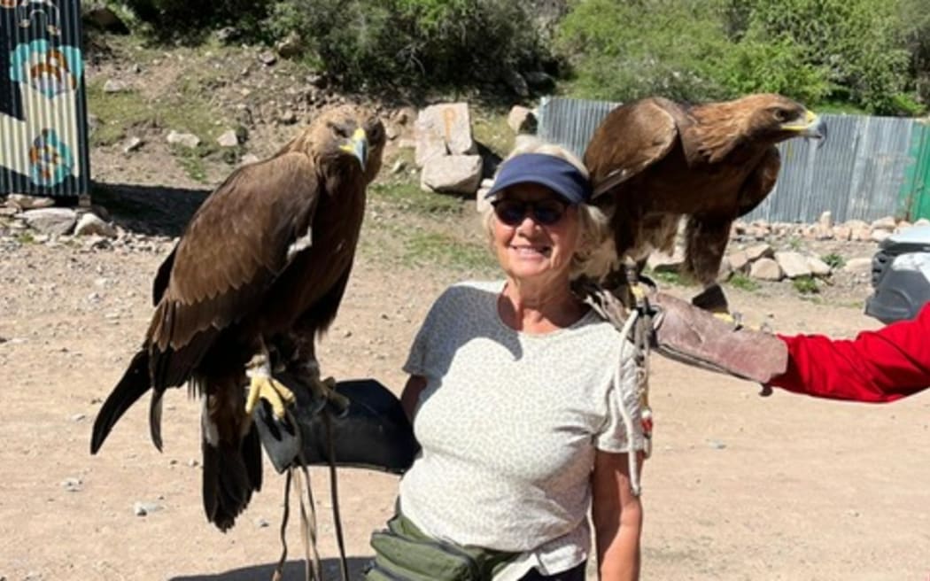 Frida Harper is wearing a blue cap and green bag. She is standing on some dusty ground in front of what looks like a rocky cliff hidden by scrub and bushes. She is holding a golden eagle on her right hand.