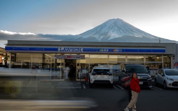 This photo taken on January 1, 2024 shows a tourist posing in front of a convenience store with Mount Fuji in the background, in the town of Fujikawaguchiko, Yamanashi prefecture. A huge black barrier to block Mount Fuji from view will be installed in a popular photo spot by Japanese authorities exasperated by crowds of badly behaved foreign tourists, it was reported on April 26, 2024. (Photo by Philip FONG / AFP)