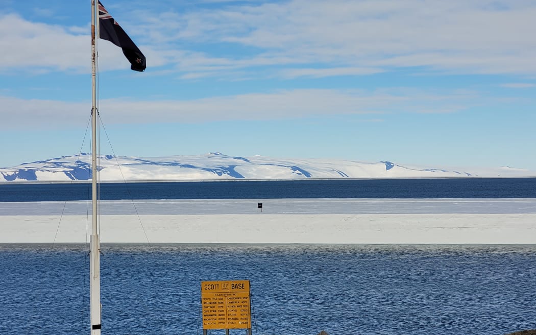 A wide shot of an open body of water with a large flat ice floe floating in the middle of it, with a flag visible on the icefloe. In the background are snow-draped hills, and in the foreground is a rocky shore with a New Zealand flag flying, and a yellow sign that reads 'Scott Base'.