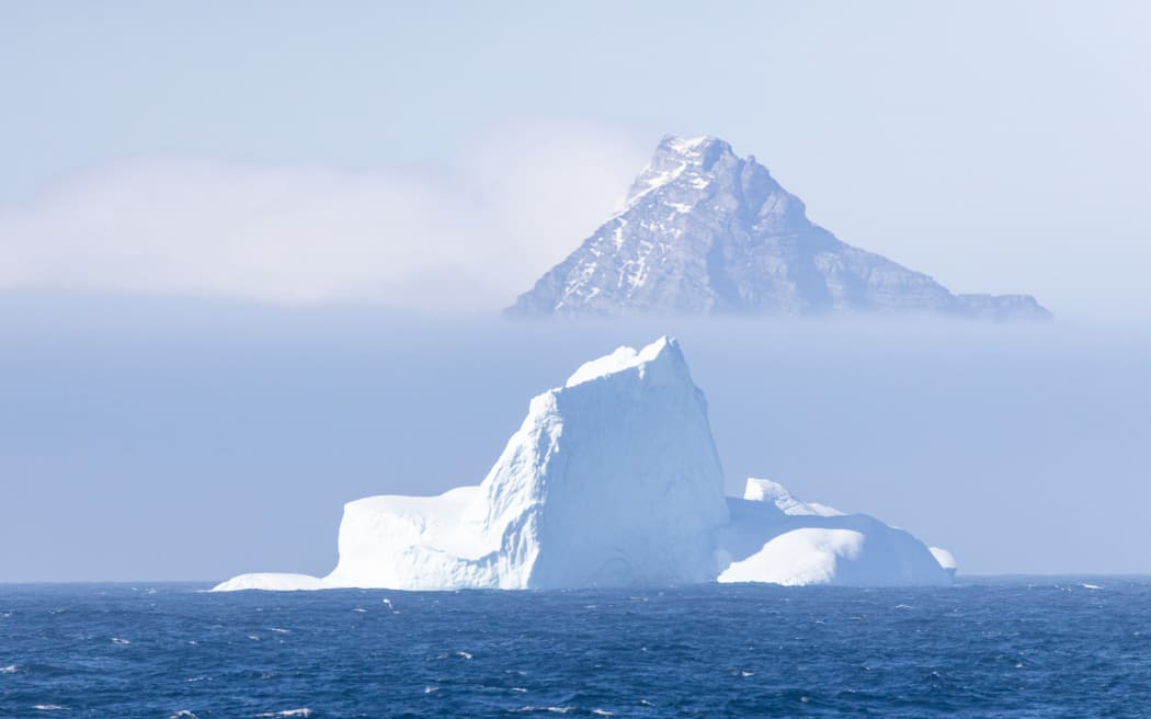 A pointy rocky peak dotted with snow peeks above a layer of dense sea fog, with a large triangular iceberg floating on dark choppy sea in the foreground.