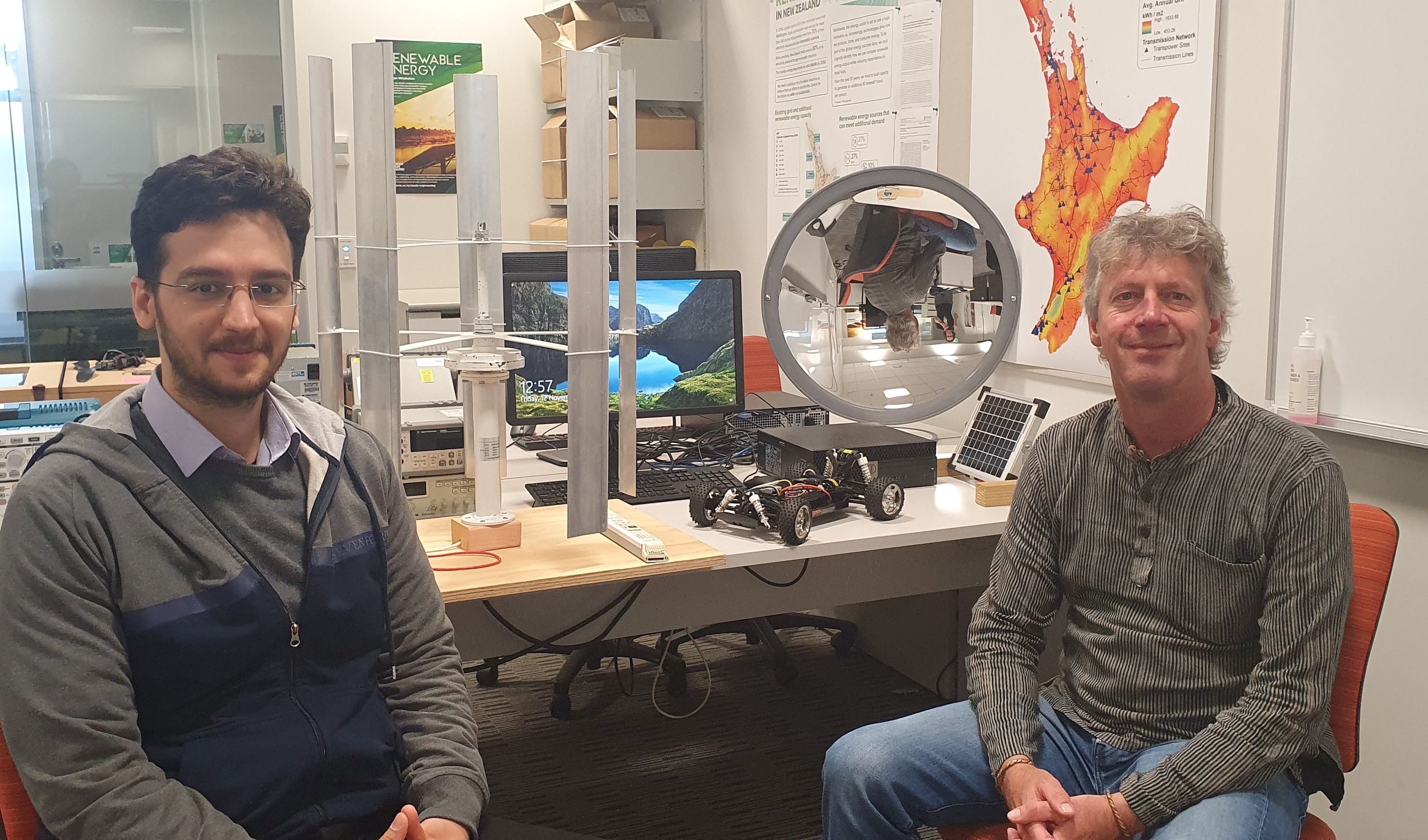 Soheil Mohseni and Alan Brent work with sustainable energy systems. There are mini versions of a vertical wind turbine and a solar concentrator on the desk behind them.