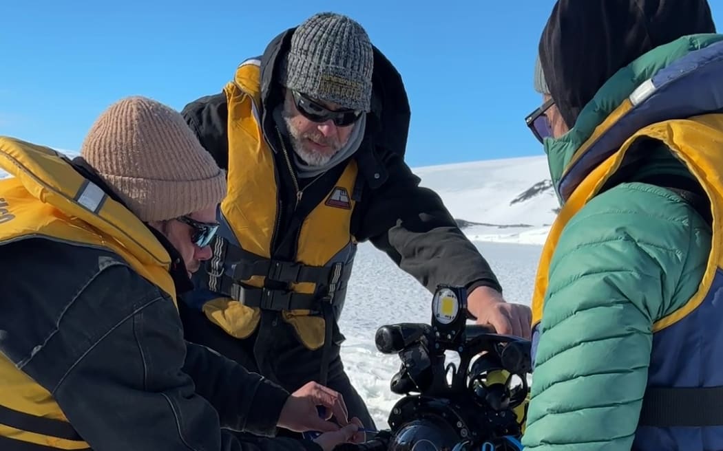 Three people wearing beanies, sunglasses, warm puffer jackets and lifejackets kneel around a camera robot. They are on a white expanse of sea ice beneath a bright blue sky.