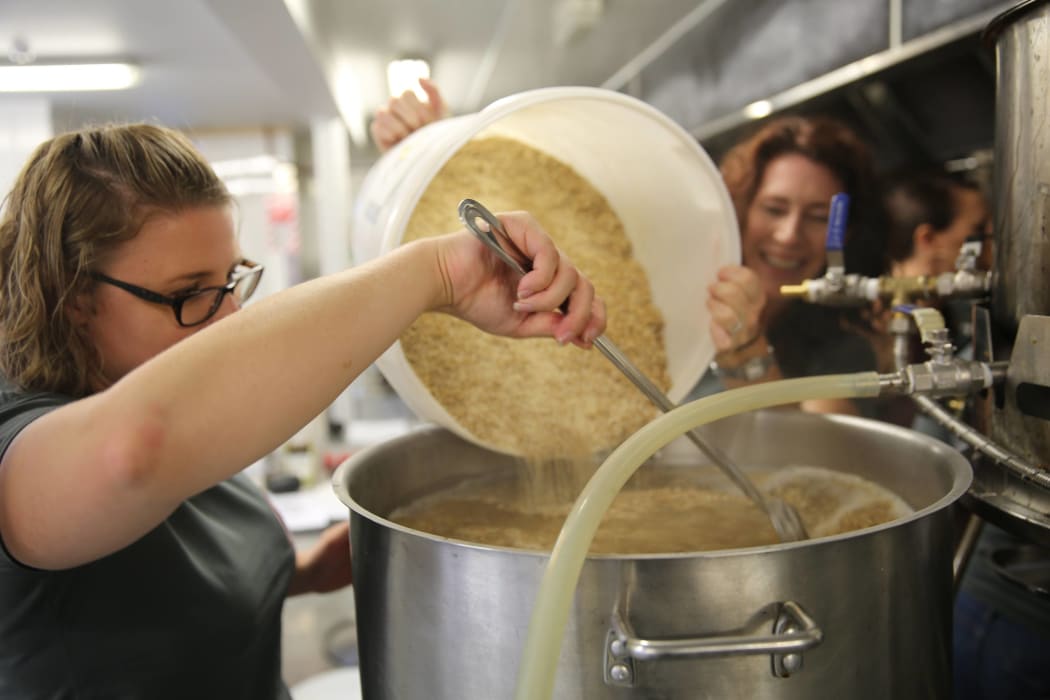 Two women pour grains from a bucket into a stainless steel tub.