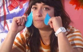 A dark haired woman in a striped t-shirt holds blue cut-out tears on her cheeks.