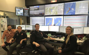 Four people on office chairs smile at the camera while sitting in front of an array of six computer monitors and ten large screens showing maps and data.
