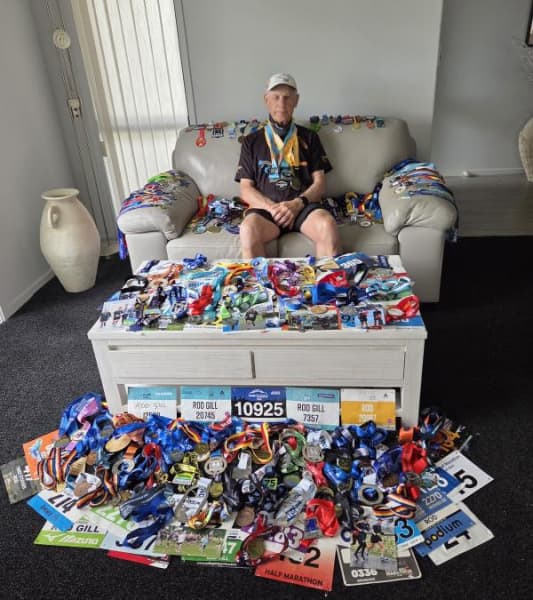 Rod Gill sits on a chair with piles of competitors medals in front of him.