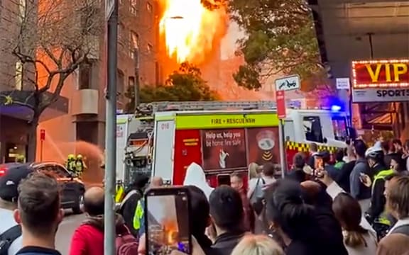 This screen grab from a UGC video taken and posted by Grant Ozolins on the Twitter account @grantozolins on May 25, 2023 shows people watching a fire in central Sydney. More than 100 firefighters battled towering flames and thick smoke from a seven-storey blaze in central Sydney on May 25 that was spreading to other buildings. (Photo by Grant OZOLINS / Twitter/@grantozolins / AFP) / -----EDITORS NOTE --- RESTRICTED TO EDITORIAL USE - MANDATORY CREDIT "AFP PHOTO / Twitter account @grantozolins/ Grant Ozolins  " - NO MARKETING - NO ADVERTISING CAMPAIGNS - DISTRIBUTED AS A SERVICE TO CLIENTS