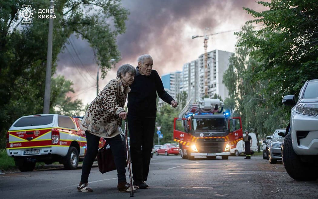 This handout photograph taken and released by the State Emergency Service of Ukraine on July 4, 2025, shows an elderly couple crossing a street as smoke rises in the background at a site of a strike following a mass Russian drone and missile attack on the Ukraine's capital Kyiv, amid the Russian invasion of Ukraine. Russia launched its largest-ever drone and missile attack on Ukraine overnight on July 4 just hours after a telephone call between US and Russian presidents ended without any breakthrough. (Photo by Handout / State Emergency Service of Ukraine / AFP) / RESTRICTED TO EDITORIAL USE - MANDATORY CREDIT "AFP PHOTO / STATE EMERGENCY SERVICE OF UKRAINE / HANDOUT" - NO MARKETING NO ADVERTISING CAMPAIGNS - DISTRIBUTED AS A SERVICE TO CLIENTS