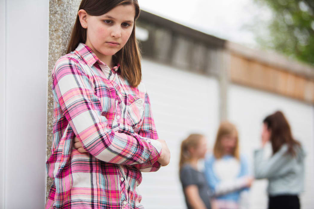 A photo of a sad looking teenage girl in the foreground. Behind her is a group of other girls talking
