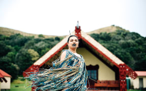 Ruth stands out the front of a marae, her clothes blowing in the wind.