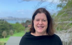 Joan is stood to the side of a tree. It has no leaves which suggests the photo was taken in the winter. Her shoulder length hair is also giving the appearance of a small amount of wind. You can see the sea in the distant background behind her.