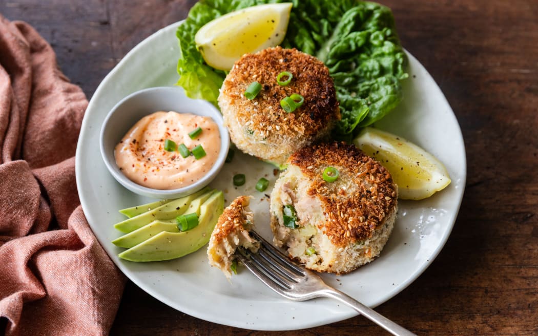 A plate containing two green curry potato and tuna fish cakes, accompanied with sliced avocado, lemon wedges and lettuce.