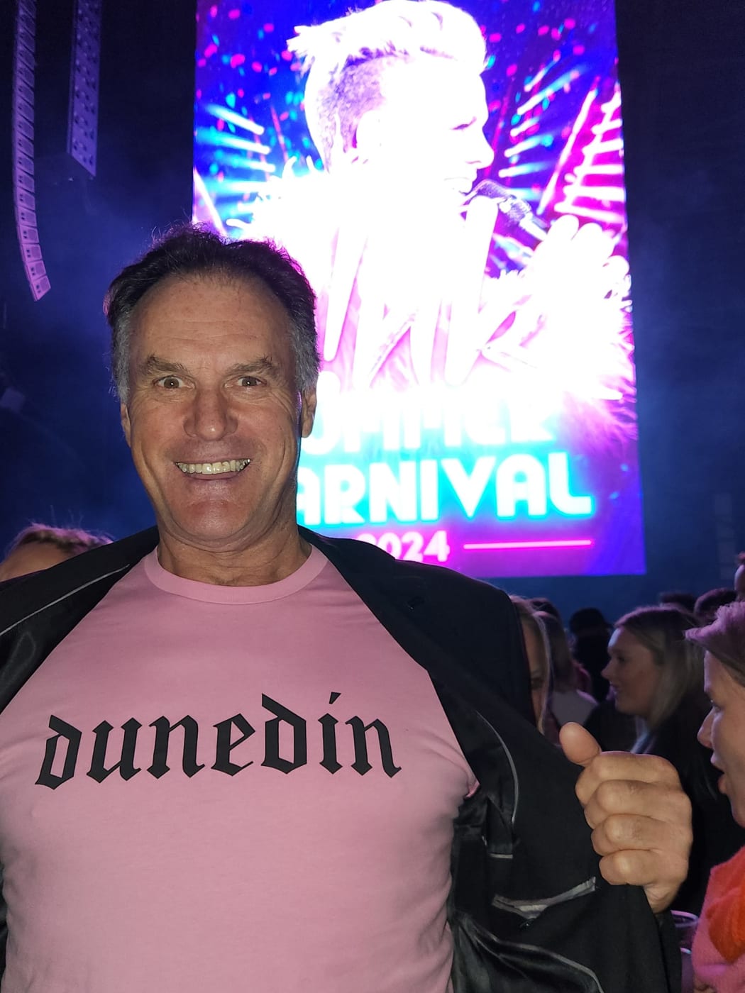 Jules Radich smiles broadly at the camera. He holds his jacket open to show a pink tshirt with the gothic logo "DUNEDIN". He is standing in the mosh of the Pink concert. There are large bright screens behind him, and hundreds of people.