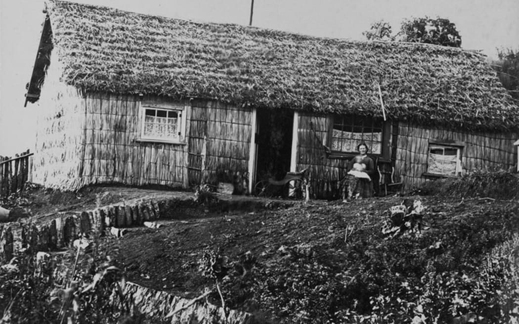 View of a mother and infant sitting outside a raupō house in Taranaki, photographed 1875–1885, by William Andrews Collis of New Plymouth. A baby’s phaeton may be seen in the doorway