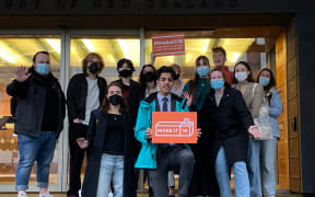 Group of young people outside court building in Auckland, one holding an orange placard that reads 'Make it 16'