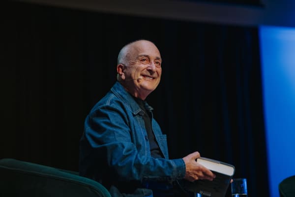 Tony Robinson, a balding man wearing a denim shirt, smiles onstage while holding a book.